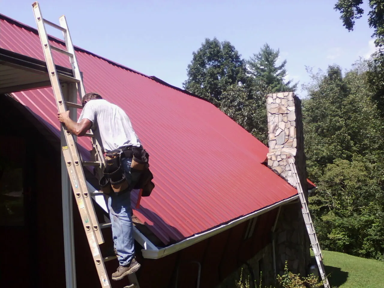 Roofer on a ladder beside a red metal roof and stone chimney — Soffit & Fascia Repair by Roofing Brothers in Ijamsville